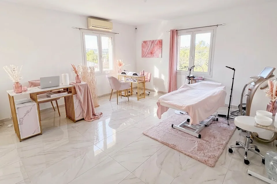 The Alicia Beauty treatment room, empty and full of light: white marble floor, a pink treatment table on a rug, a gold desk and pink chairs, two windows looking out on the hills.