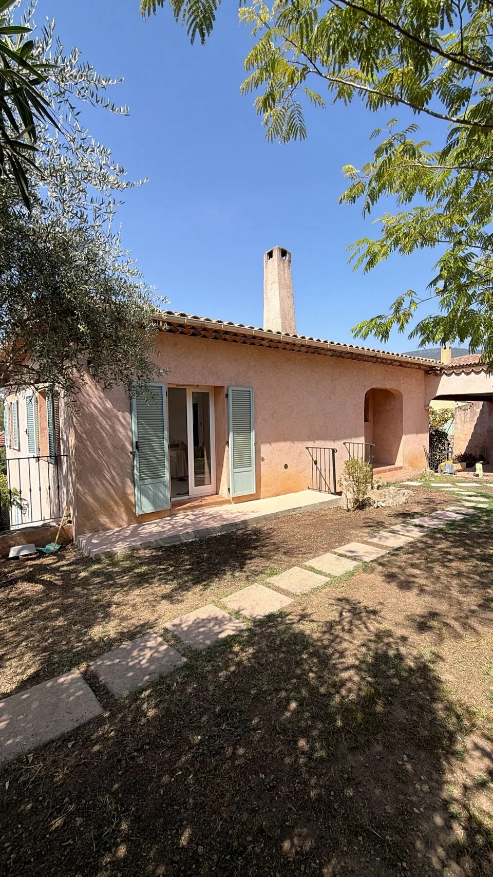 The front of the cabinet in Grasse: a Provençal house in pink render, grey-green shutters and an open French window, an arched porch and stepping stones across the garden, under a blue sky.