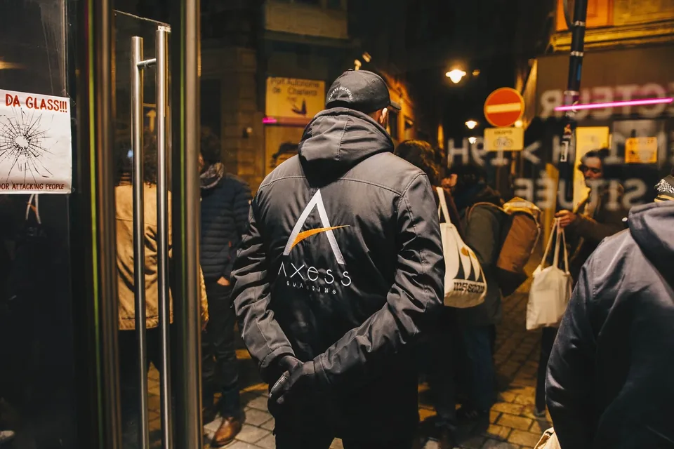 Seen from behind, an Axess Guarding officer in an embroidered cap and jacket, hands behind his back, watches over the entrance of a Charleroi venue on a busy night.