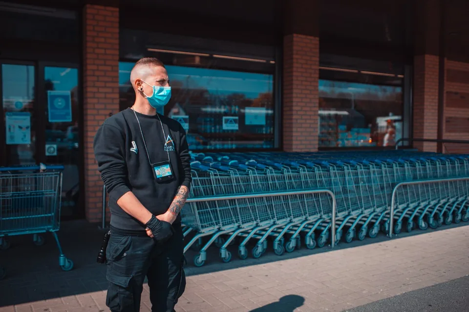 An Axess Guarding officer in a mask and gloves, an officer’s card around the neck, outside a supermarket entrance and its rows of trolleys, in April 2020.