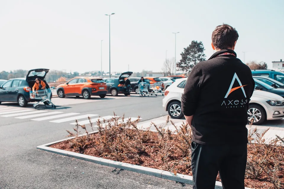 Seen from behind, an Axess Guarding officer watches over a supermarket car park where shoppers load their groceries, in April 2020.