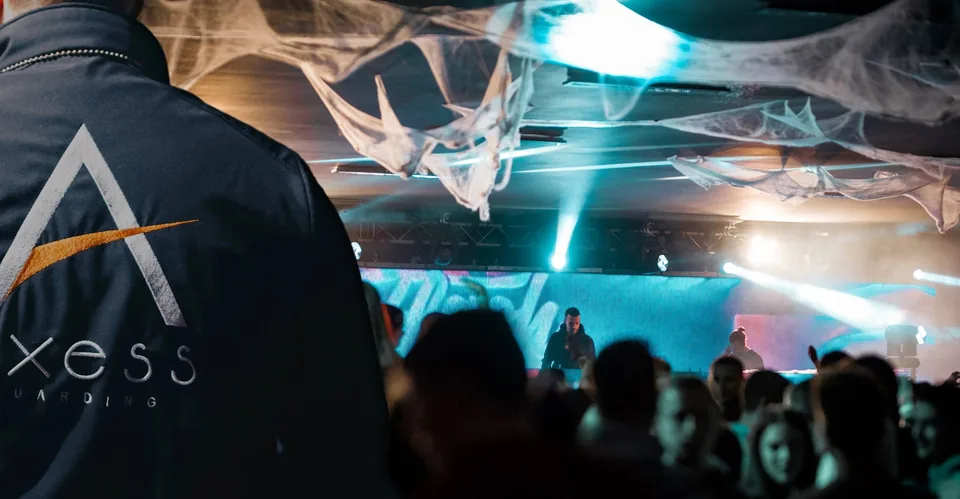 In the foreground, the embroidered back of an Axess Guarding officer; ahead of him, a GLOW’IN party crowd facing the DJ beneath cobweb decorations.