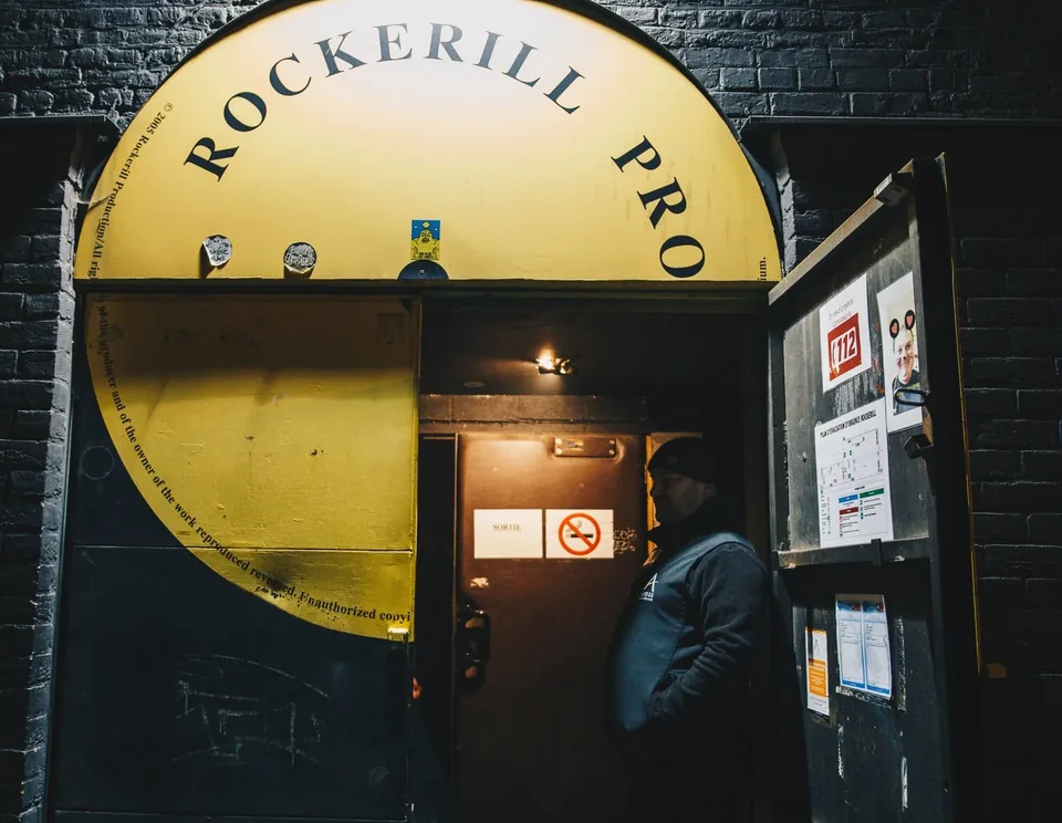 The entrance of Rockerill, in Charleroi, beneath its yellow arch; an Axess Guarding officer in a beanie stands in the doorway.