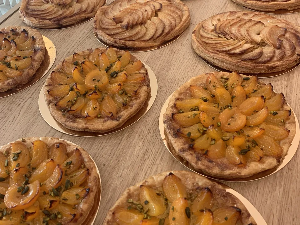 Des tartes aux abricots et pistaches tout juste sorties du four, à côté de tartes aux pommes en rosace, sur un plan de bois.