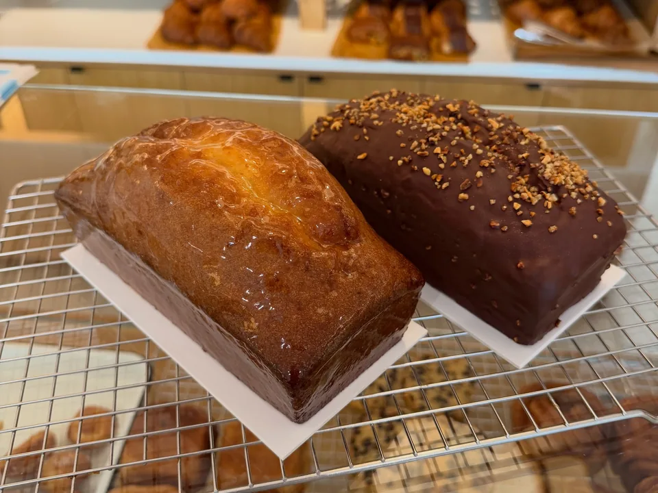 Un cake au citron glacé et un cake marbré au chocolat sur une grille, devant les étagères de viennoiseries de la boutique.