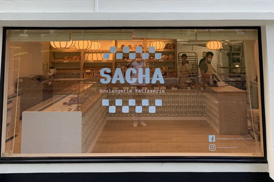 La vitrine de Sacha sur la place d’Ohain : le nom SACHA et « Boulangerie Pâtisserie » entre deux rangées de carrés bleu clair, et derrière la vitre la boutique toute neuve, ses comptoirs et ses suspensions en papier.