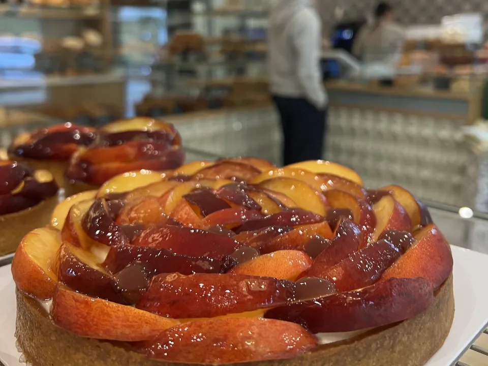 Une tarte aux pêches en rosace, nappée, au premier plan dans la boutique ; derrière, flous, les comptoirs et les suspensions.