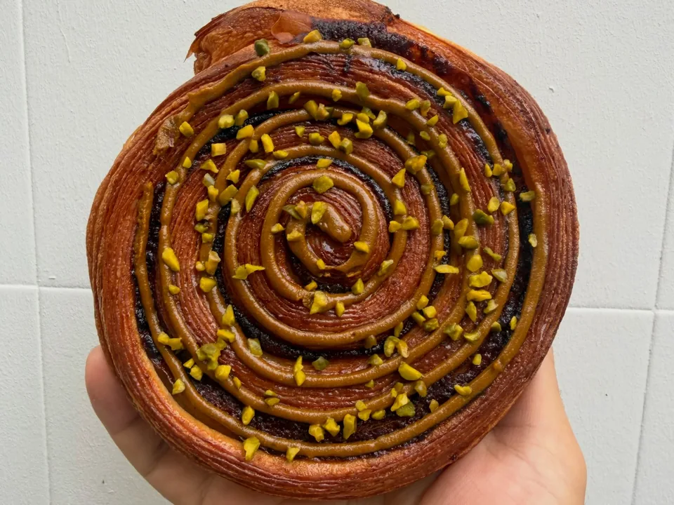 Un feuilleté roulé praliné-pistache parsemé d’éclats de pistache, tenu à la main devant un mur carrelé blanc.