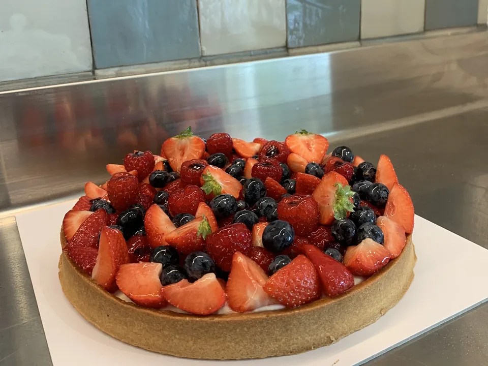 Une tarte aux fraises, framboises et myrtilles sur un plan de travail en inox, devant le carrelage bleu et crème de l’atelier.