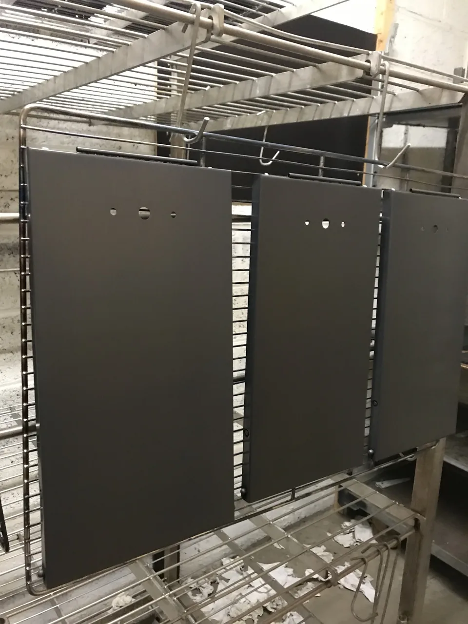 Trois plaques d’acier percées, d’un noir mat uniforme, suspendues sur un rack inox à l’atelier.