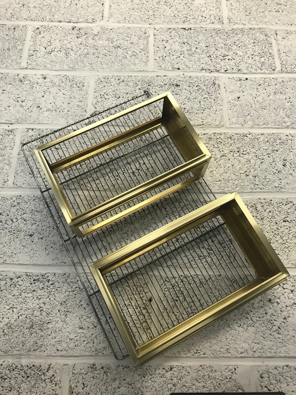 Deux cadres rectangulaires en laiton doré à fond grillagé, posés sur une grille contre un mur de blocs béton.