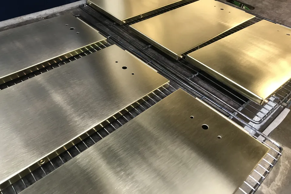 Six brushed golden brass panels, folded and drilled, lying flat on drying racks in the workshop.