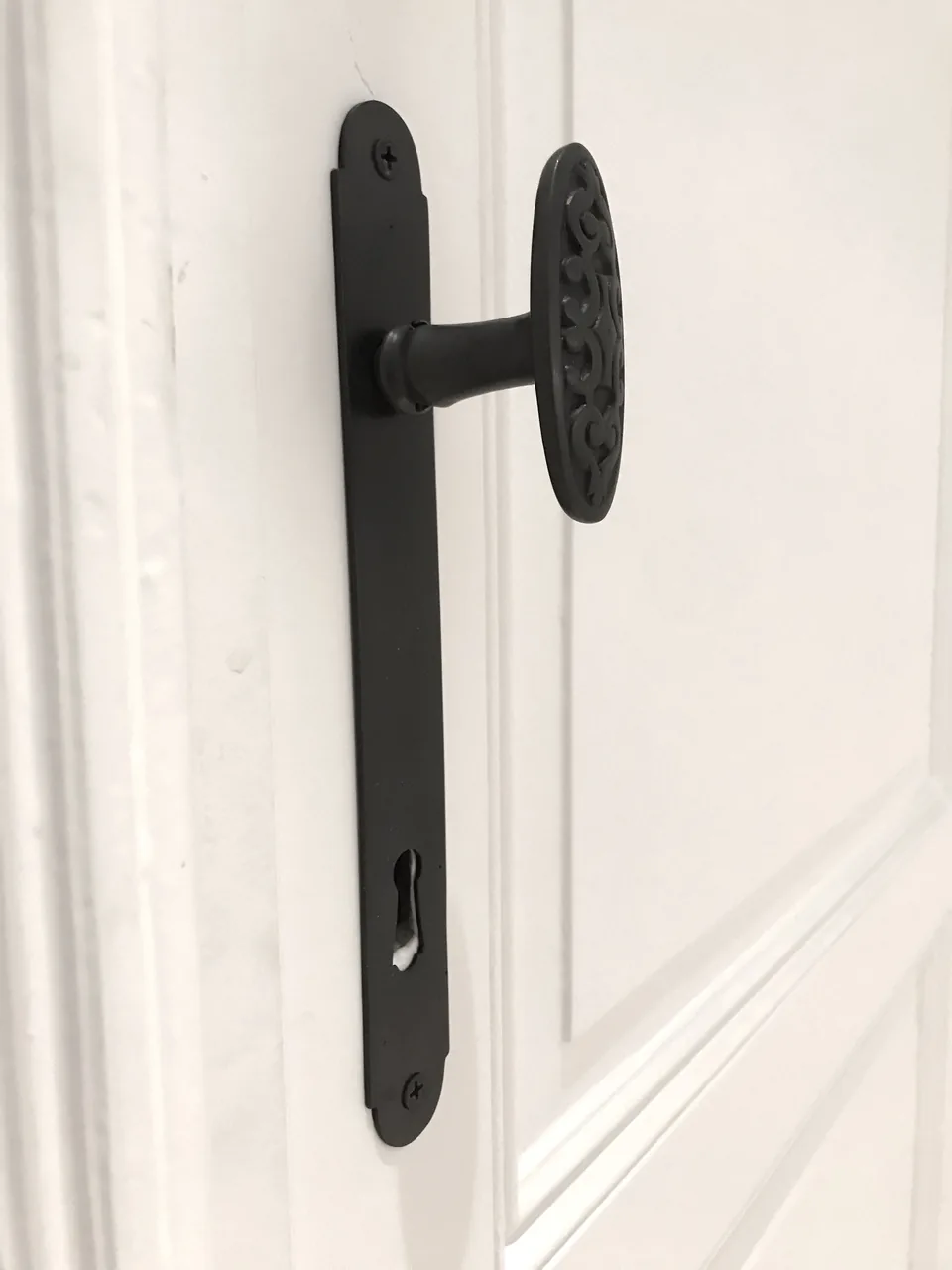 Une béquille à longue plaque et un bouton ovale ouvragé en métal noir mat, sur une porte blanche.