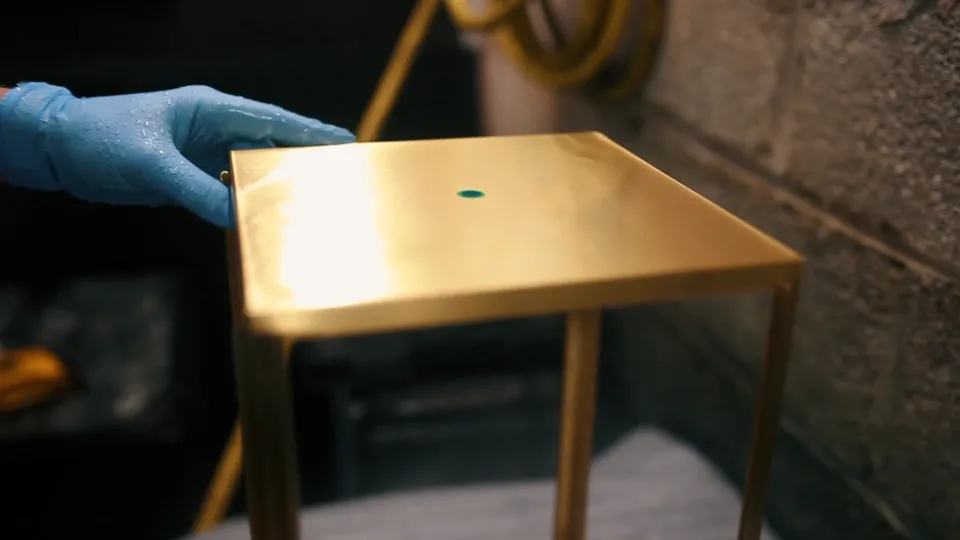A blue-gloved hand sets down a small golden brass table, a green dot in the middle of its top, against a block wall.