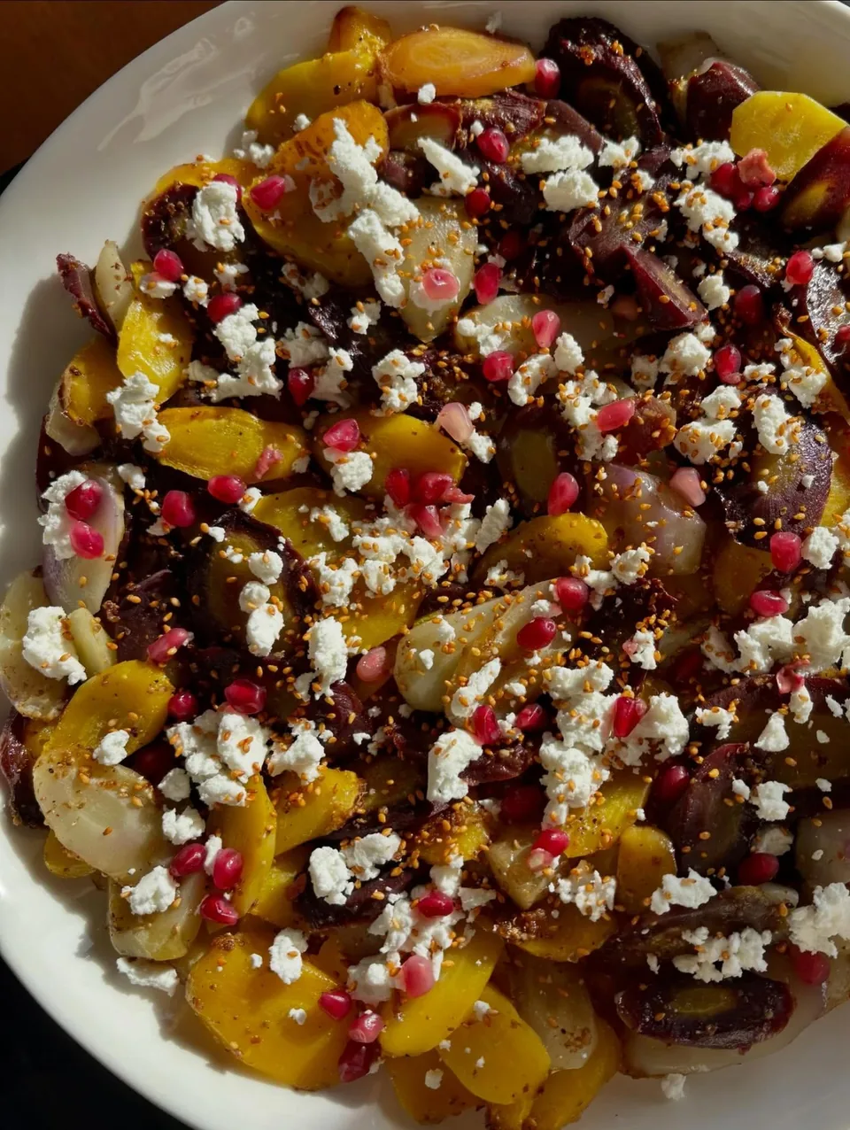 Un plat du comptoir traiteur vu de dessus : légumes rôtis jaunes et violets, feta émiettée, graines de grenade et graines grillées.
