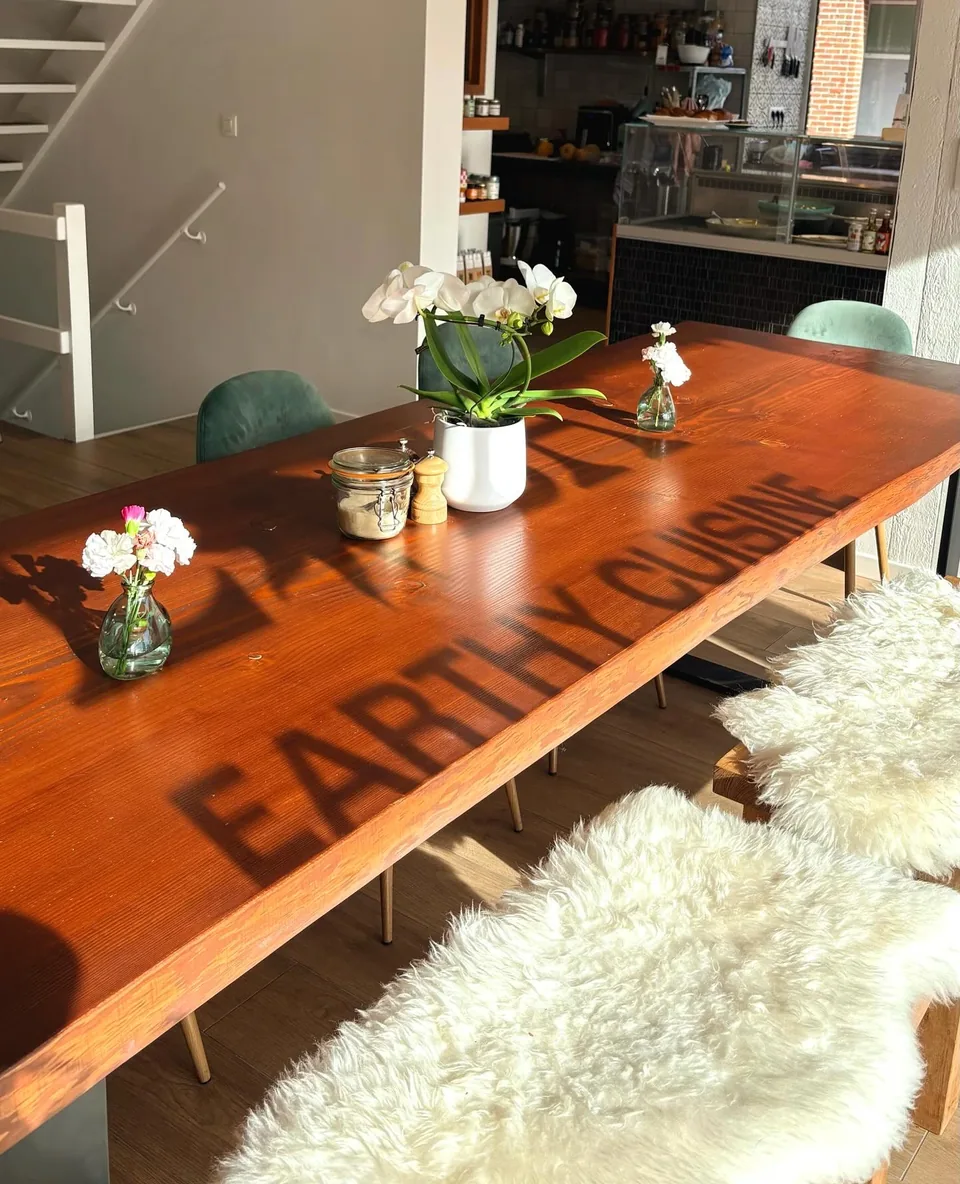 La grande table en bois de la salle au soleil, des fleurs blanches en petits vases, des peaux de mouton sur les chaises ; l’ombre des lettres « Earthy cuisine » de la vitrine se dessine sur le bois.