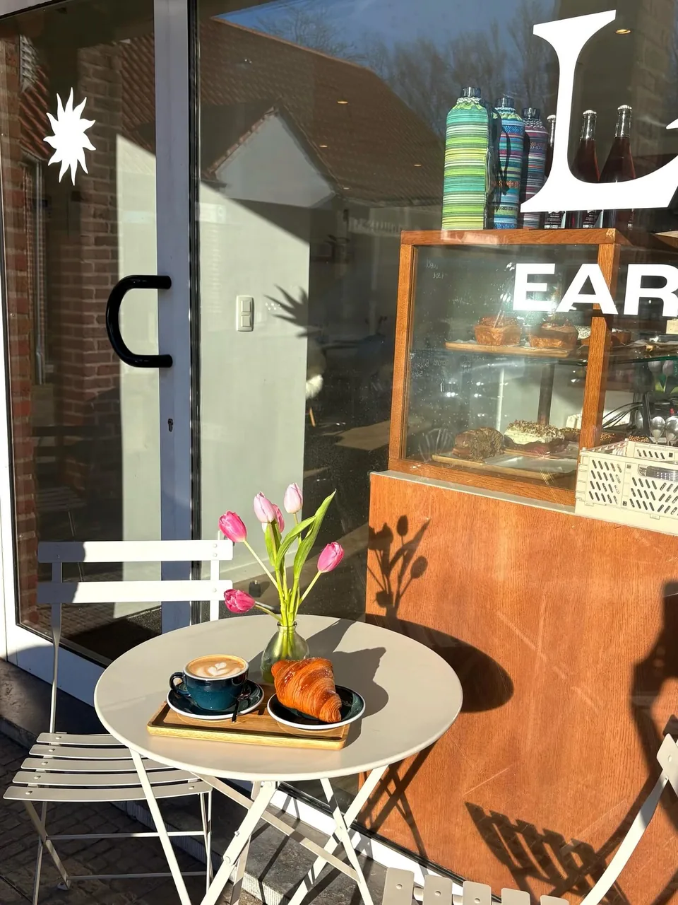 Sur la terrasse, une table ronde blanche avec un cappuccino, un croissant et des tulipes roses, devant la vitrine au nom de Loba et le comptoir des gâteaux.