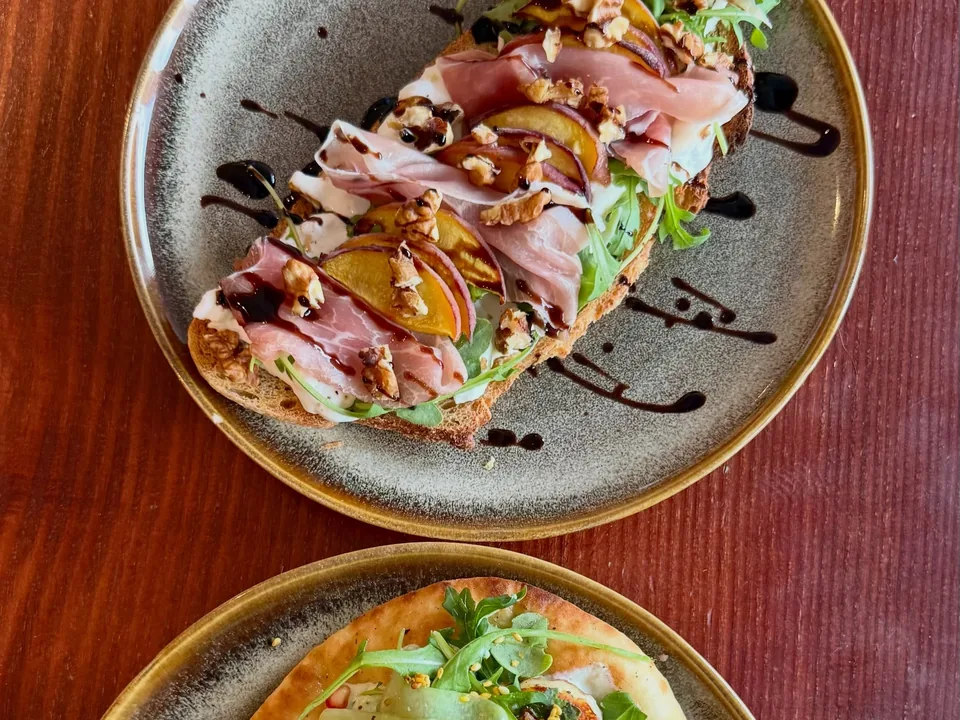 Les nouvelles tartines du brunch sur deux assiettes en grès : l’une au jambon cru, pêche, noix et roquette, l’autre sur pain plat aux rubans de concombre et à la grenade.