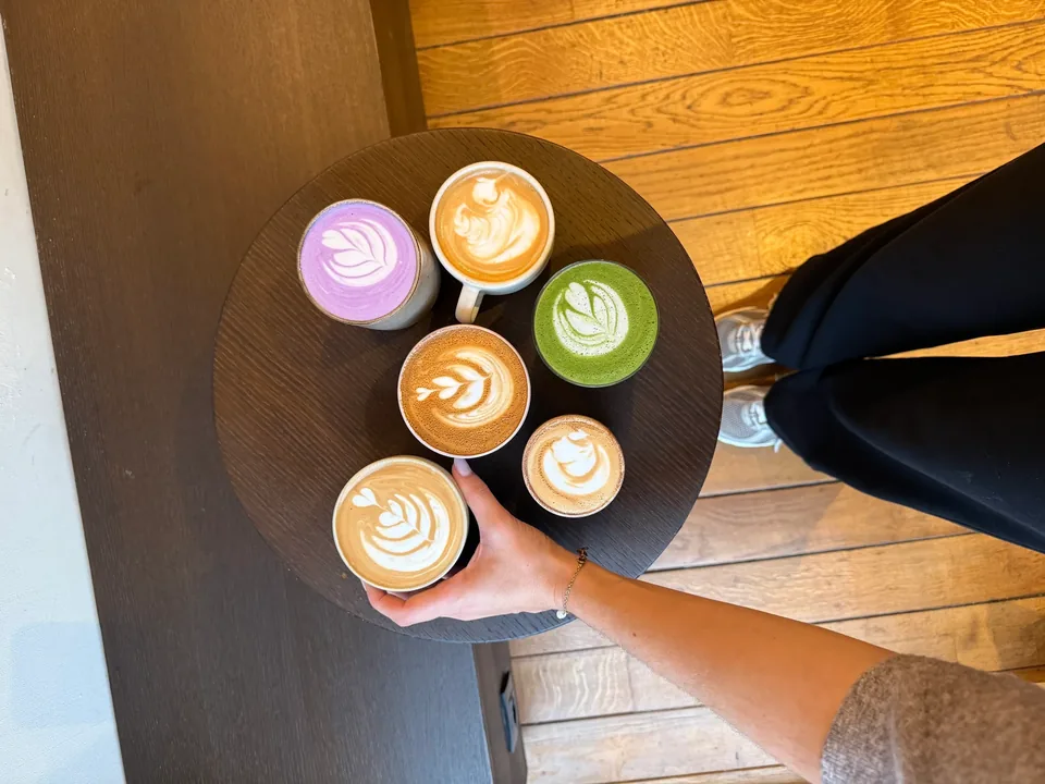 Vu d’en haut, un plateau rond de six cafés au lait dessinés, dont un ube violet et un matcha vert.