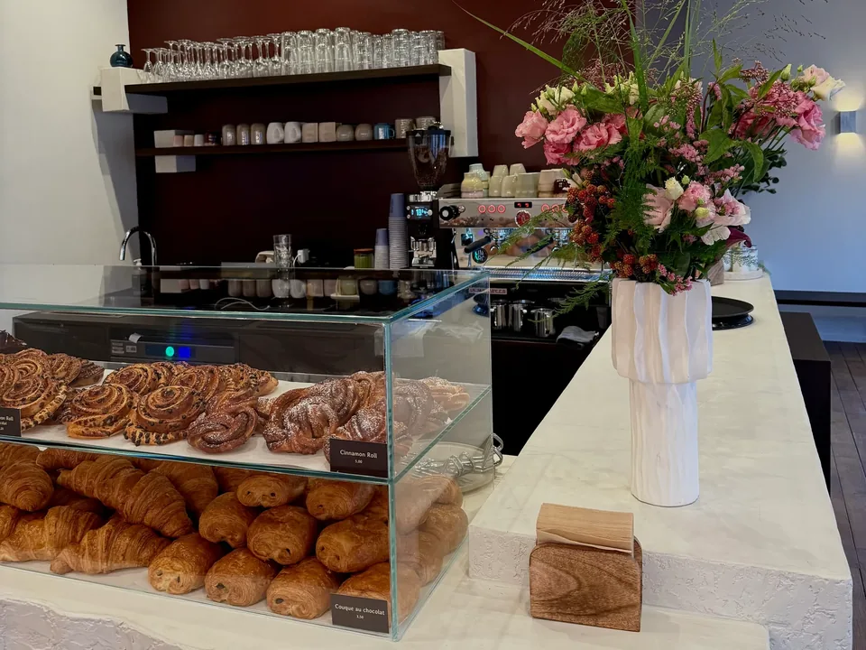 Le comptoir blanc : une vitrine de rolls et de croissants, un grand bouquet de fleurs, et derrière, la machine à espresso et les étagères de tasses.