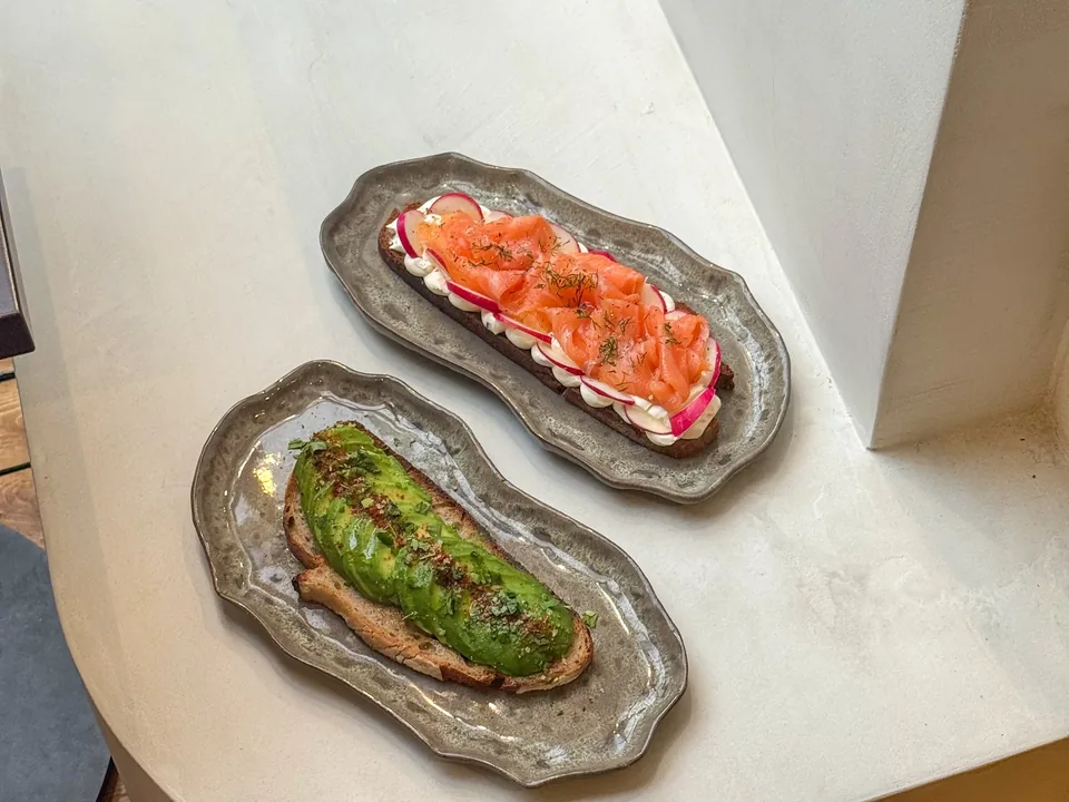 Deux tartines sur des plats en grès, l’une au saumon fumé, l’autre à l’avocat, posées sur la table blanche.