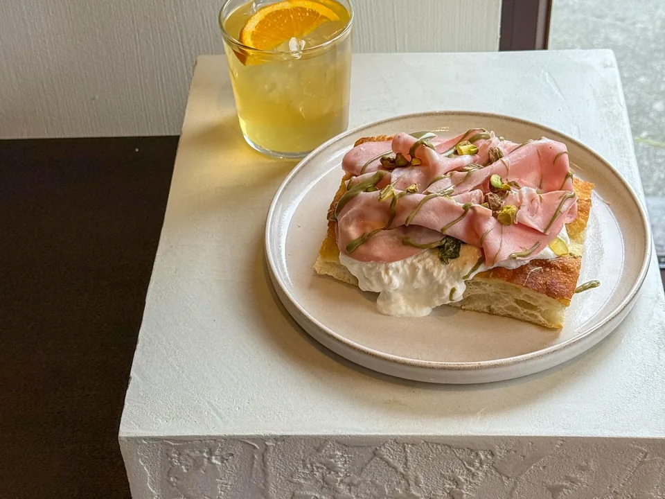 Une part de focaccia garnie de burrata, de fines tranches de charcuterie et de pistaches, un thé glacé à l’orange, sur un bloc blanc près de la vitre.