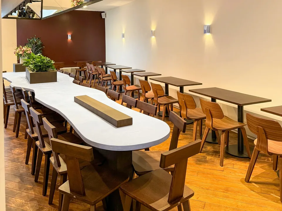 The wavy white table and its wooden chairs under the blue ceiling light, small tables lined up along a wall lit by sconces.