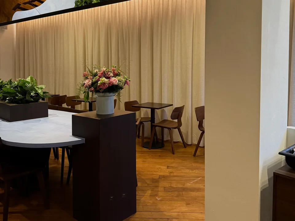A corner of the room along a tall pale curtain: the white table, a bunch of pink flowers and green plants under a dark ceiling box.