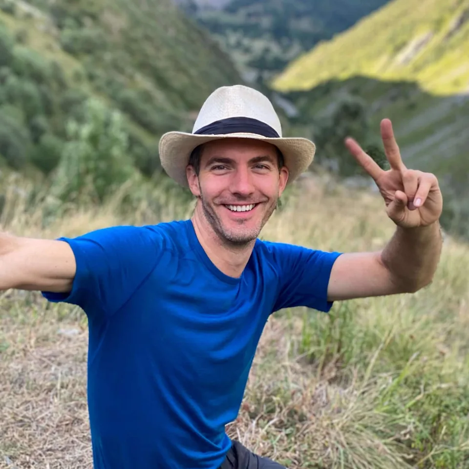 Alexis Bertrand, smiling, in a straw hat and a blue T-shirt, makes a victory sign in front of a green valley.