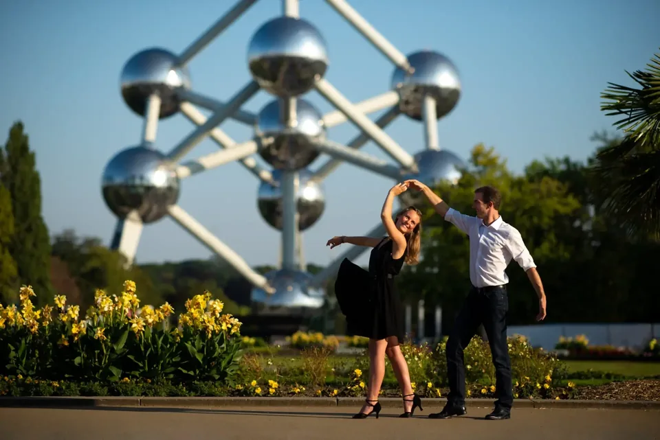 A couple dances in front of the Atomium, between beds of yellow flowers: he in a white shirt, she in a black dress, arms raised.