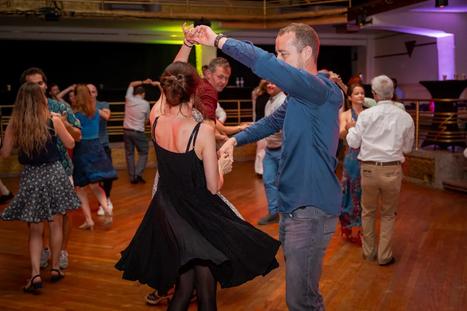 Couples dance rock on a large wooden floor; in front, a dancer in a blue shirt spins his partner in a black dress.
