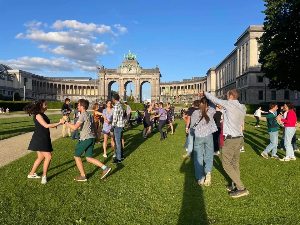 Couples dance rock on the lawn of the Cinquantenaire park, the arch and its colonnades behind, under a blue sky.