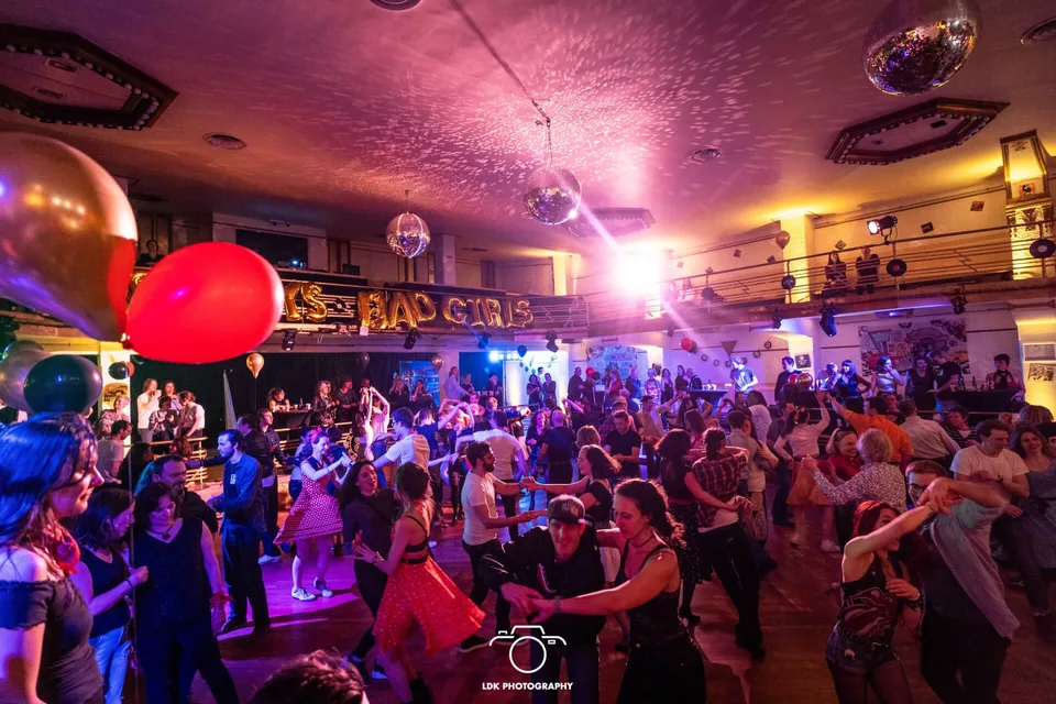 A packed hall under mirror balls and balloons: dozens of couples dance, a « Bad Girls » sign on the balcony.