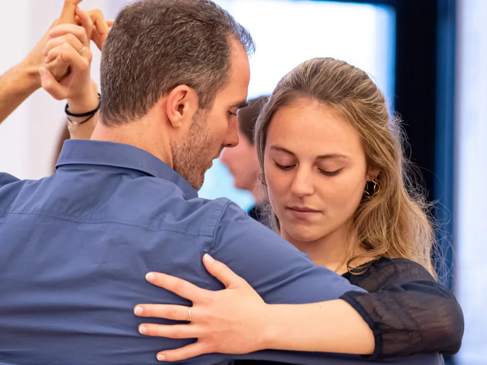 A Slow Rapide couple in a close hold, eyes lowered: he in a blue shirt, she in a black blouse, by a window.