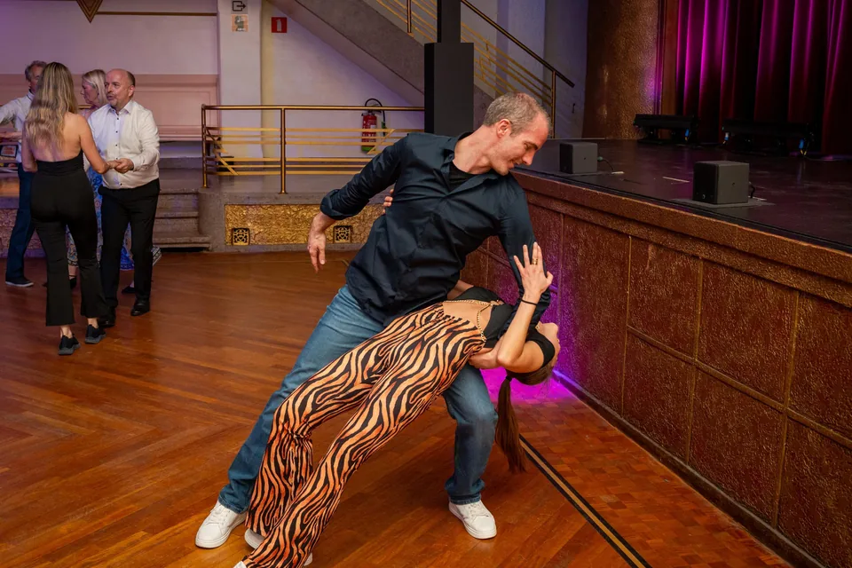 A dancer in a black shirt dips his partner, in tiger-striped trousers, down to the floor, in front of a stage with purple curtains.