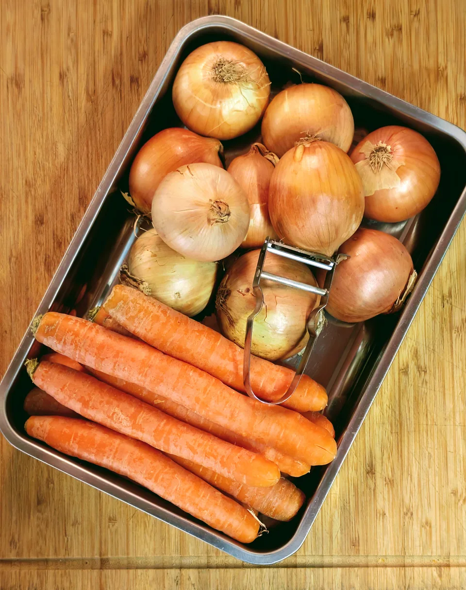 Un bac en inox rempli d’oignons et de carottes, un économe posé dessus, sur une planche en bambou de la cuisine du Club House.