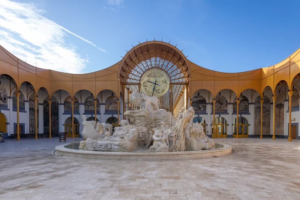 The entrance of Pairi Daiza, the Gallery of the Worlds: a paved courtyard with golden arcades, a great clock under an arched glass roof and a fountain sculpted with animals.
