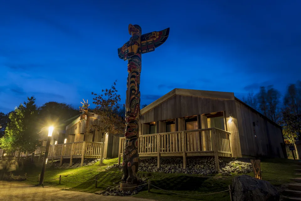A carved totem pole and the wooden lodges of the Last Frontier, lit at nightfall.