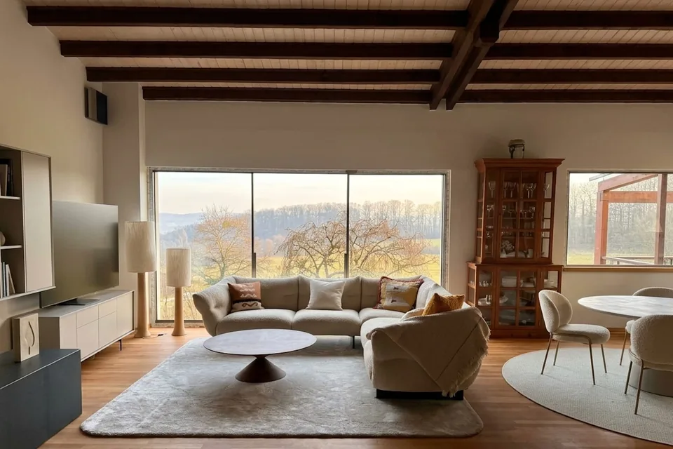 The Catherine project: a living room beneath exposed beams, a three-panel panoramic bay with slim profiles opening onto the winter countryside, a bouclé sofa and a wooden floor.