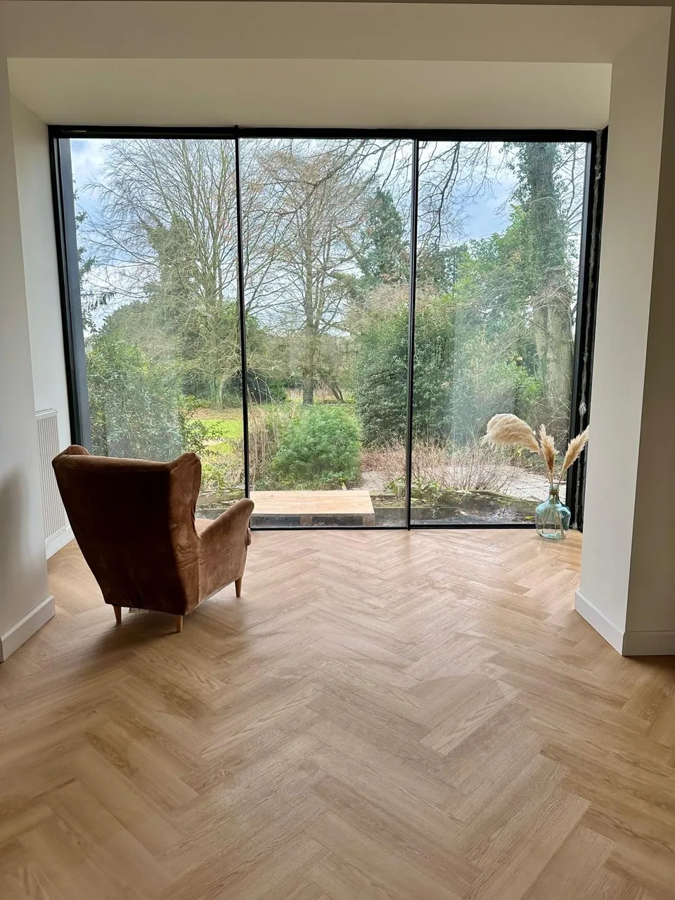 The Ellie project: a floor-to-ceiling three-panel sliding bay with very slim black profiles, opening onto a wooded garden, a builder’s pallet still outside; a leather armchair and a herringbone floor.