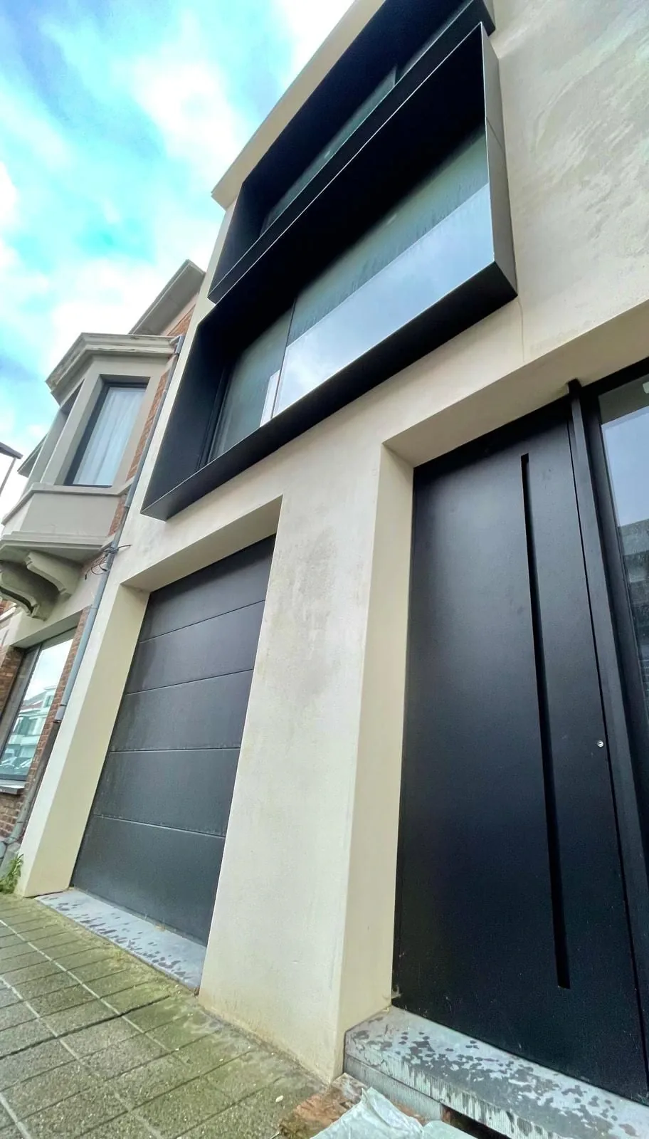 The Françoise project, as the work was finishing: a rendered façade, a tall black pivot door, a black garage door and, upstairs, a window in a projecting black frame.