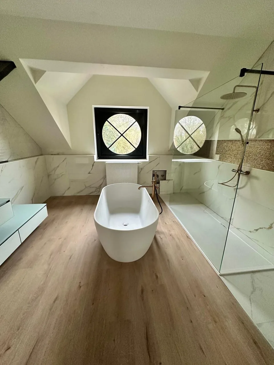 The Sophie project: an attic bathroom, a freestanding bath, two round windows with black cross bars, a glass walk-in shower and marble-effect tiles.